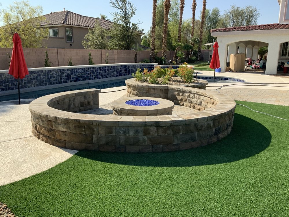 A circular stone wall surrounds a firepit with blue glass stones next to a pool